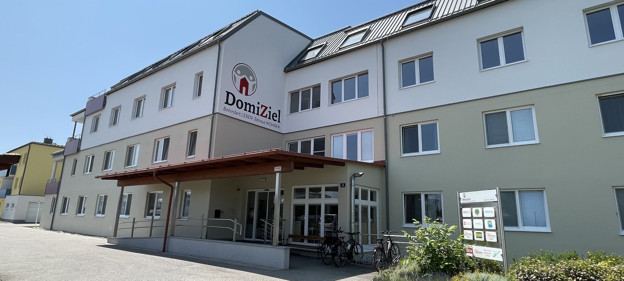 Dreistöckiges Wohnhaus mit einem Vordach beim Eingangsbereich und der Aufschrift DomiZiel