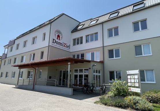 Wohnhaus mit Erdgeschoss zwei Obergeschossen und einem Dachgeschoss und dem Schriftzug DomiZiel Behindert LEBEN. Betreut WOHNEN. seitlich auf der Fassade