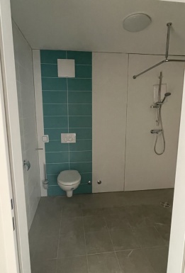 Einblick in ein Badezimmer mit weißem hängenden WC auf teilweise hellblauer Fliesenwand einem grauen Fliesenboden und einem Duschbereich rechts