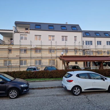4 stöckiges weißes Gebäude mit grauem Schrägdach zum Teil eingerüstet mit Autos davor