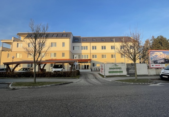 4 stöckiges helles Wohnhaus mit grauem Schrägdach Baustellenzaun und Bäumen davor