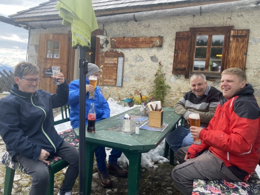 Eine Frau und drei Männer sitzen lachend vor einer Berghütte und trinken Bier und Cola