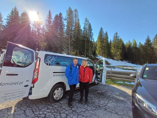 Zwei Männer stehen vor dem DomiZiel Bus auf einem Parkplatz und im Hintergrund sieht man die Bäume und die Sonne
