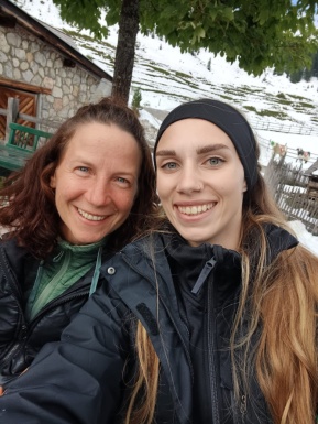 Zwei Frauen lachend mit einer schneebedeckten Wiese und einer Hütte im Hintergrund
