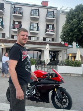 Mann vor Hotel steht vor einem Motorrad und lächelt