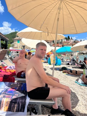 Zwei Männer sitzen auf einer Liege im Strand unter einem Sonnenschirm