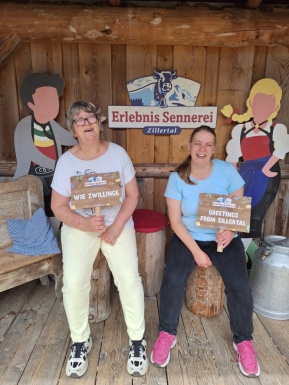 Zwei Frauen sitzen auf Holzhockern vor einem Schild mit der Aufschrift Erlebnis Sennerei