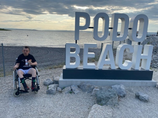 Mann im Rollstuhl neben Podo Beach Skulptur am Strand vom See