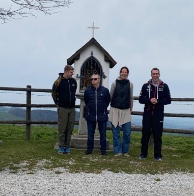 Drei Männer und eine Frau stehen vor einem Andachtsbildstock und einem Berggipfel