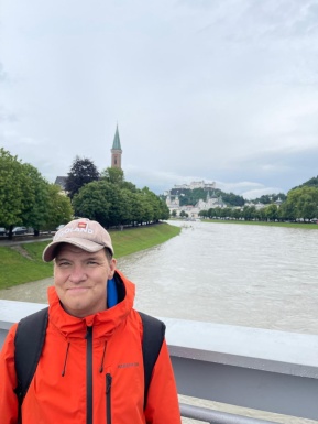 Mann mit Kappe und Regenjacke auf Brücke mit Wasser im Hintergrund