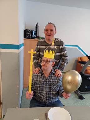 Beim Tisch sitzender Mann mit Krone, goldenem Ballon und Zepter in den Händen und dahinter legt ihm ein lächelnder Mann die Hände auf die Schultern