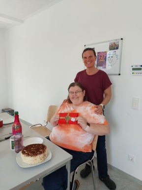 Frau steht hinter einer sitzenden Frau die Geburtstag hat und legt ihr die H&auml;nde auf die Schultern