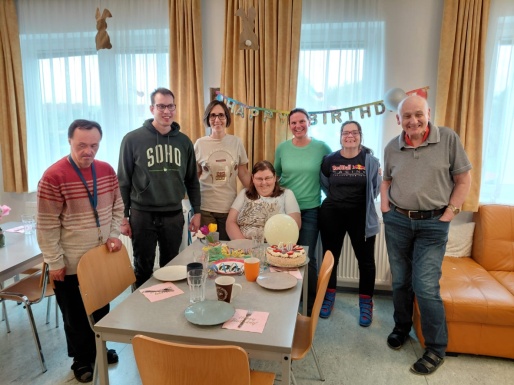 Gruppe von Menschen stehen um einen Tisch mit Torte, Ballon und Geschirr wo eine Frau sitzt