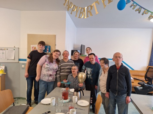 Gruppe von Menschen steht um einen mit Geschirr gedeckten Tisch wo ein Mann sitzt mit einem goldenen Ballon in der Hand