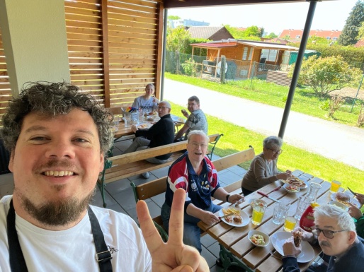 Mann l&auml;chelt und zeigt 2 Finger in die H&ouml;he w&auml;hrend andere Leute im Hintergrund auf der Terrasse essen