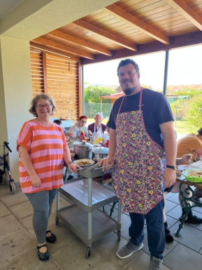 Eine Frau mit gestreiftem Tshirt und ein Mann mit bunter Sch&uuml;rze stehen auf der Terrasse und l&auml;cheln