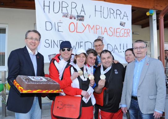 Menschengruppe teilweise mit Medaillen umgehängt und ein Mann mit einer großen Torte in den Händen stehen vor einem Plakat mit Glückwünschen für die Olympiasieger