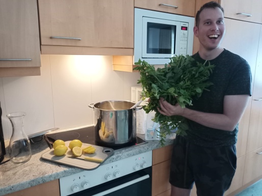 Lächelnder Mann steht in einer Küche vorm Ofen mit einem Strauß Minze in der Hand und auf dem Ofen liegt ein Brett mit Zitronen und es steht ein Topf darauf