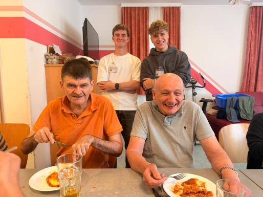 Zwei M&auml;nner sitzen beim Tisch mit Essen und zwei M&auml;nner stehen dahinter