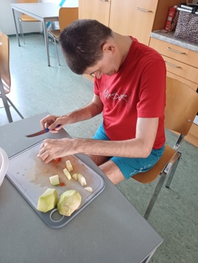 Ein Mann mit blauer kurzer Hose und einem roten T-shirt sitzt beim Tisch und schneidet Gemüse
