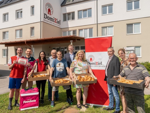 Gruppenfoto mit Menschen die Schachteln mit Backwaren und Gutscheinen in den Händen halten vor Wohnhaus mit DomiZiel Aufschrift