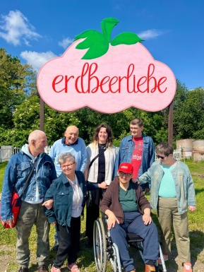 Menschen stehen gemeinsam mit einem Mann im Rollstuhl vor einem Schild mit der Aufschrift "Erdbeerliebe"